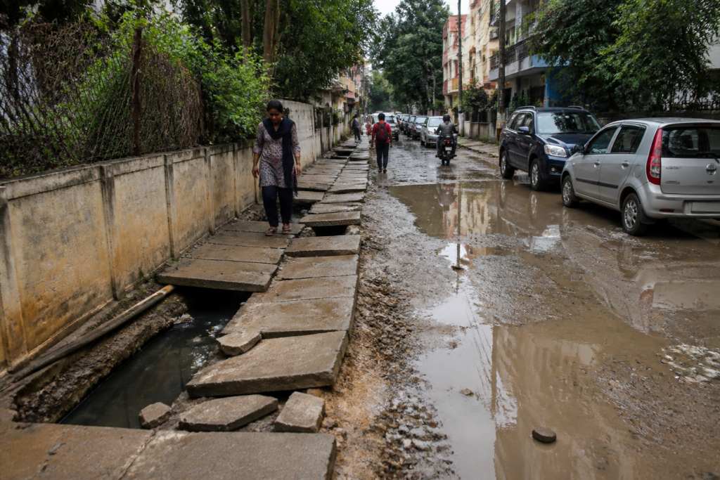 A damaged footpath beside an open drain and muddy road in a Bengaluru neighbourhood