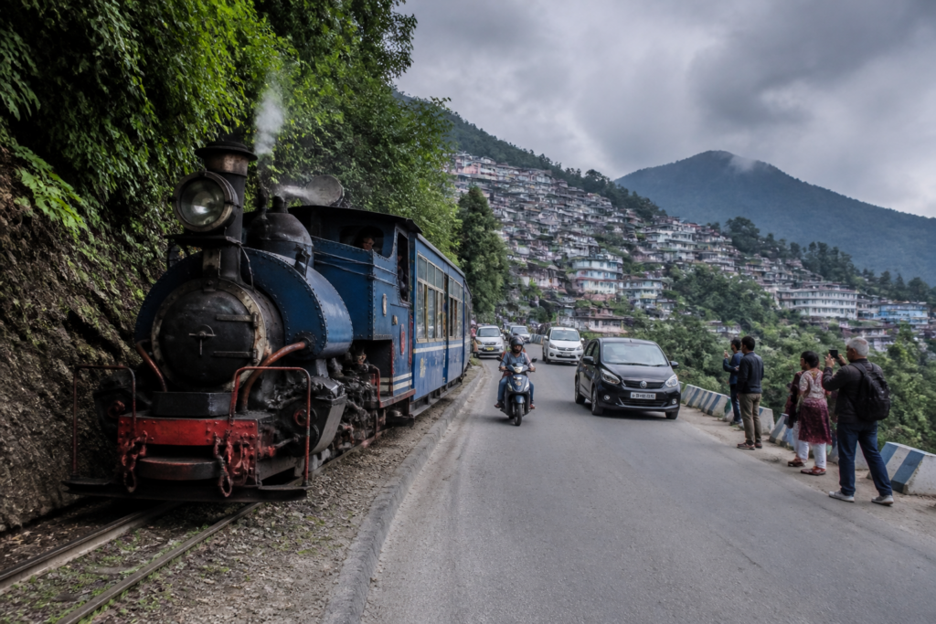 Darjeeling toy train moving beside a narrow hill road with vehicles and tourists nearby