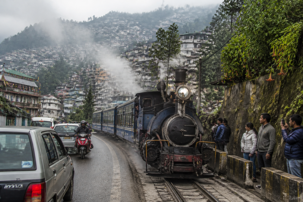 Darjeeling toy train passing beside a narrow hill road with vehicles nearby