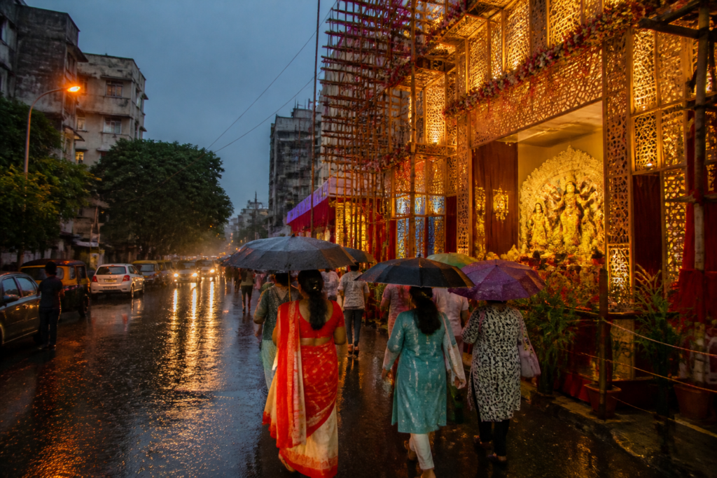 Kolkata Puja weather with rain clouds over a decorated pandal and pedestrians carrying umbrellas