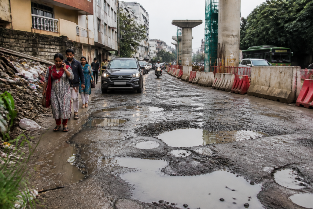 Damaged S K Garden road with potholes and construction debris beside an obstructed footpath
