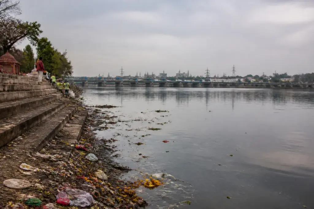 Yamuna pollution at a Delhi ghat with litter along the riverbank and urban infrastructure beyond